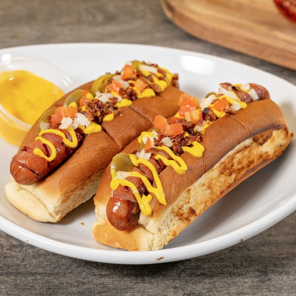 Two hot dogs with mustard, relish, diced tomatoes, and onions on a white plate.