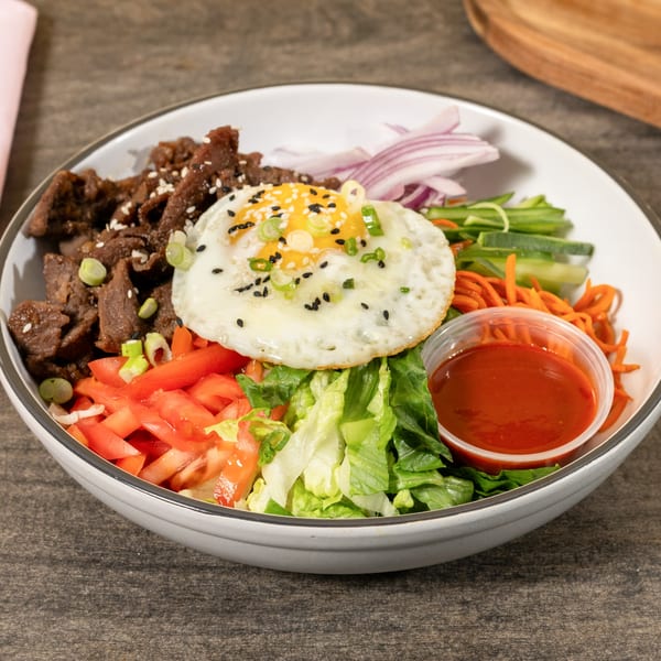 Bibimbap bowl with beef, fried egg, red onions, carrots, cucumbers, tomatoes, lettuce, and sauce.