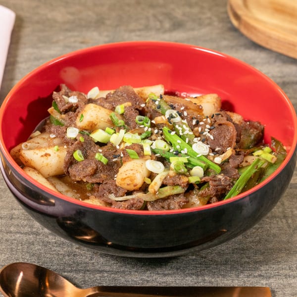 A black and red bowl filled with Asian beef and rice cakes, garnished with green onions and sesame seeds.