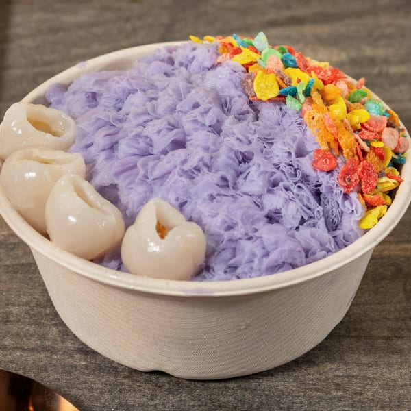 Purple shaved ice with lychee and colorful cereal in a white bowl on a wooden table.